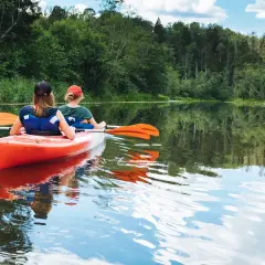 Dwie osoby płyną kajakiem po spokojnej rzece, otoczonej zielenią. Idealne miejsce na spływ kajakowy Mazury, gdzie najlepiej poczuć bliskość natury.